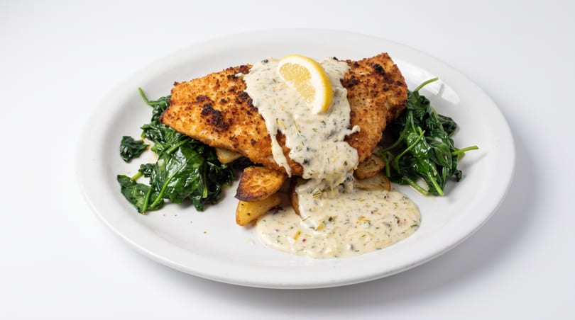 Breaded fish fillet with creamy sauce, lemon, spinach, and roasted potatoes on a white plate.