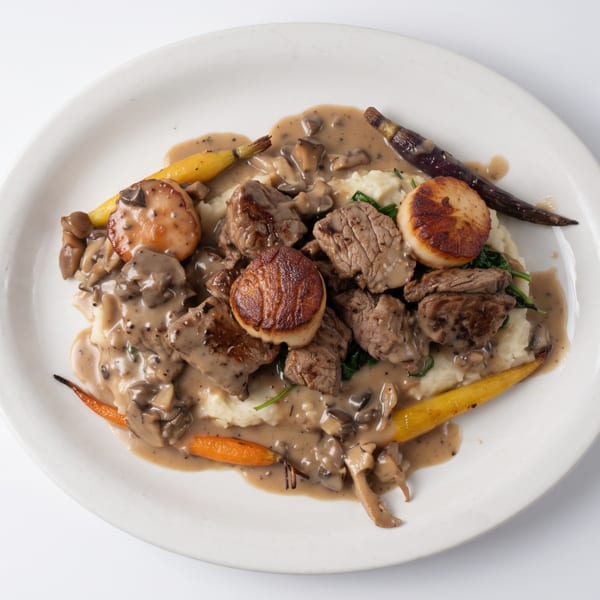 Seared scallops and beef with mushroom sauce, mashed potatoes, and carrots on a white plate.