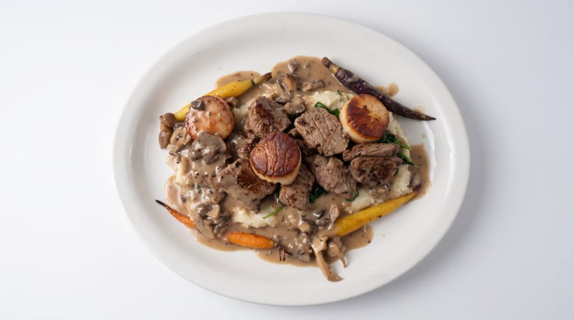 Seared scallops and beef with mushroom sauce, mashed potatoes, and carrots on a white plate.
