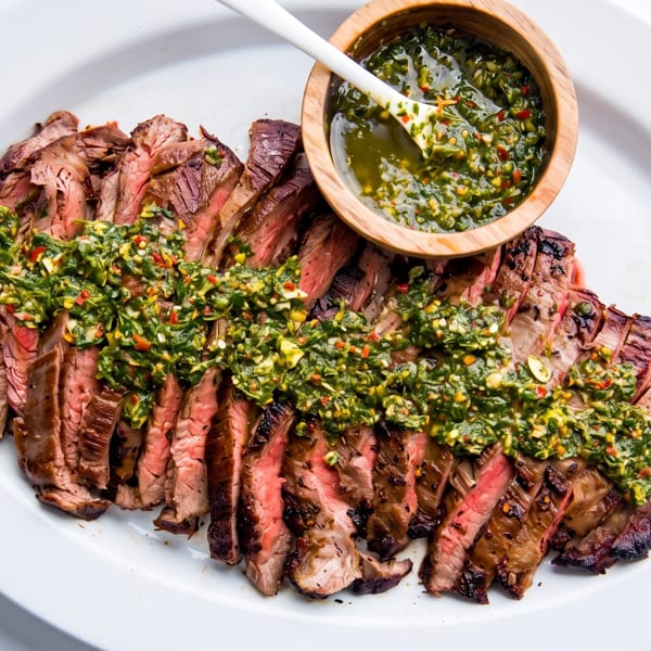 Perfectly grilled sliced steak with vibrant green chimichurri sauce on a white platter.