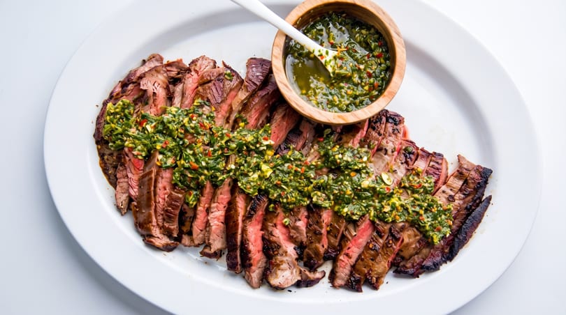 Perfectly grilled sliced steak with vibrant green chimichurri sauce on a white platter.