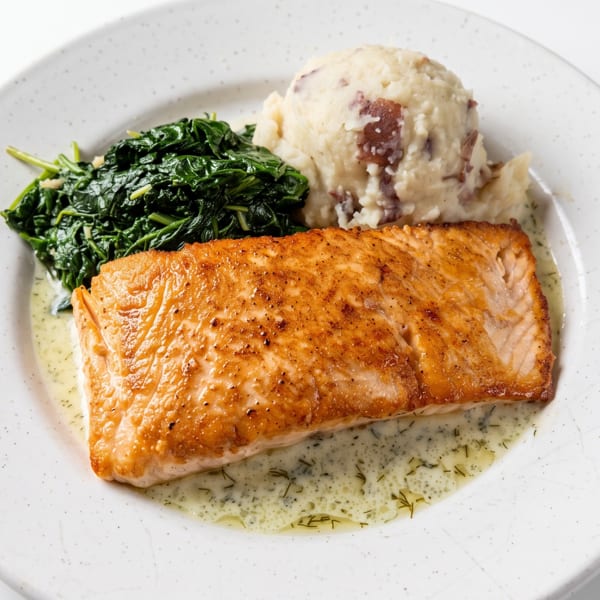 Pan-seared salmon with creamy dill sauce, sautéed spinach, and rustic mashed potatoes on a white plate.