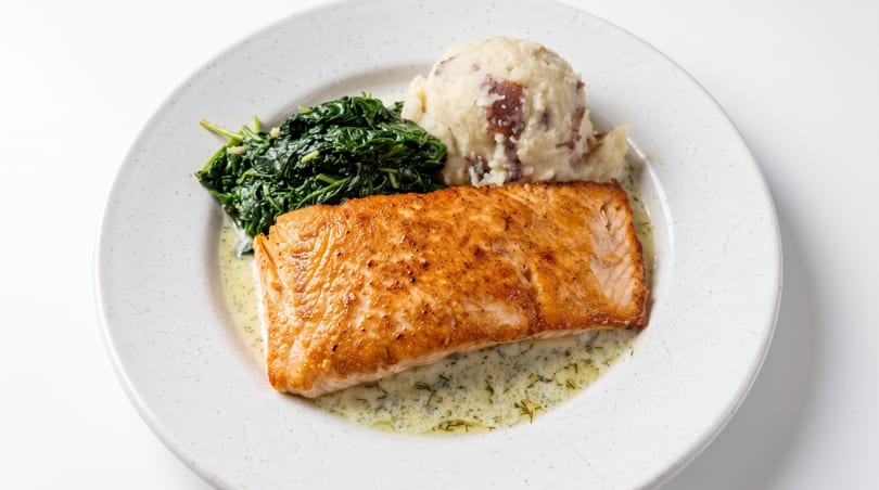Pan-seared salmon with creamy dill sauce, sautéed spinach, and rustic mashed potatoes on a white plate.