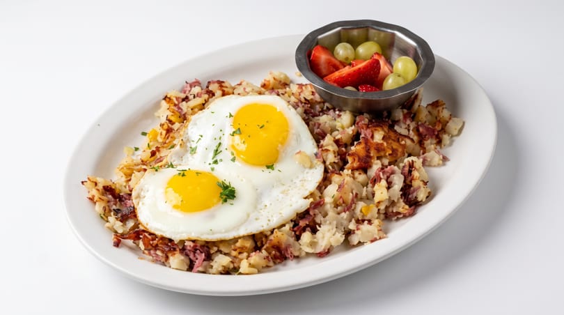 A breakfast plate featuring corned beef hash, two sunny-side-up eggs, and a fruit cup.